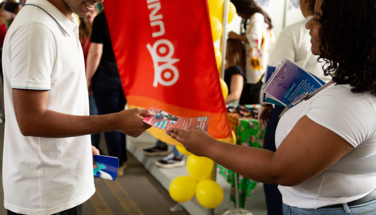 Pessoa entrega um material informativo a um estudante durante evento institucional da UNISUAM. Ao fundo, há um banner da instituição, balões amarelos e outros participantes circulando pelo espaço.