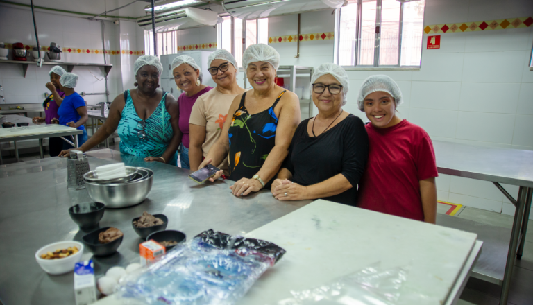 Grupo de participantes usando toucas de proteção posa para uma foto em uma cozinha industrial equipada para atividades práticas de gastronomia.