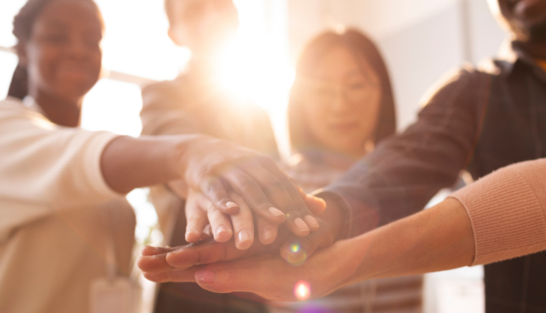 Grupo de pessoas com diferentes perfis e origens coloca as mãos umas sobre as outras, simbolizando união, colaboração e trabalho em equipe.