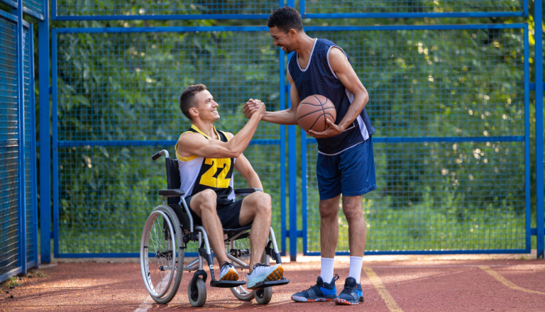 Dois homens em uma quadra esportiva ao ar livre sorriem e cumprimentam-se com um aperto de mãos. Um deles está em uma cadeira de rodas e veste uniforme de basquete; o outro segura uma bola de basquete.