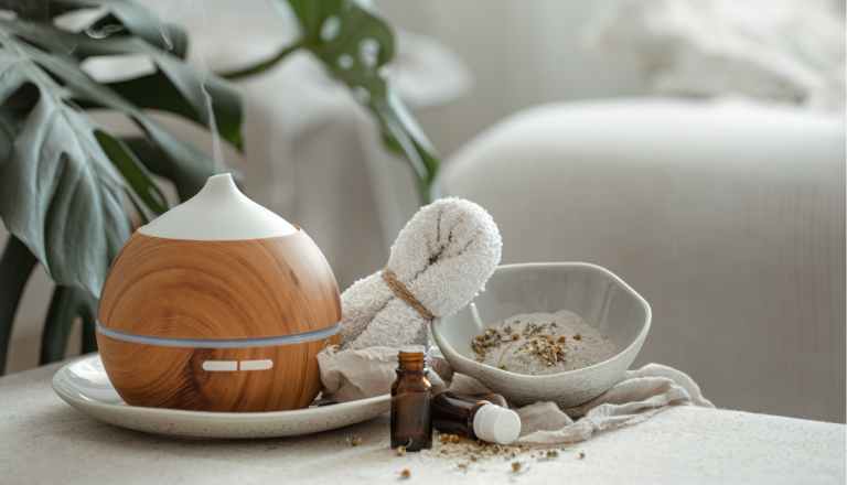 Difusor de aromas com acabamento amadeirado sobre uma bandeja branca, ao lado de uma toalha enrolada, frascos de óleo essencial e uma tigela com ingredientes naturais. Ao fundo, elementos decorativos em tons claros criam uma atmosfera aconchegante e relaxante.