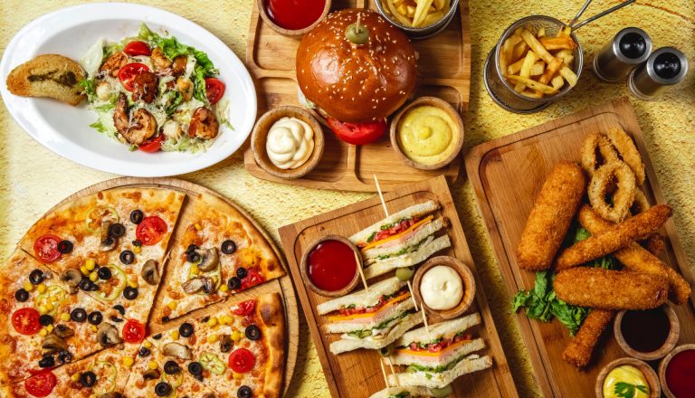 Há uma pizza de vegetais, um hambúrguer com batatas fritas, sanduíches acompanhados de molhos, uma porção de alimentos empanados com anéis de cebola, uma salada Caesar com frango grelhado e diferentes acompanhamentos, compondo uma mesa repleta de opções.