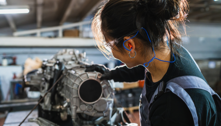 Pessoa usando óculos de proteção, protetores auriculares e luvas trabalha na manutenção de um componente mecânico em uma oficina industrial.