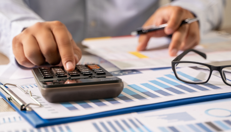 Pessoa utilizando uma calculadora enquanto analisa relatórios financeiros com gráficos e tabelas. Sobre a mesa, há documentos, uma caneta e um par de óculos.