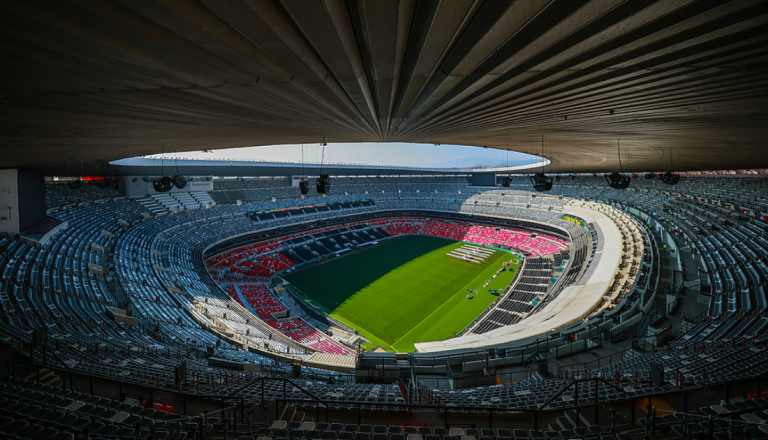 Vista panorâmica de um estádio de futebol vazio, com arquibancadas em tons de cinza e vermelho ao redor de um gramado verde iluminado pela luz natural.