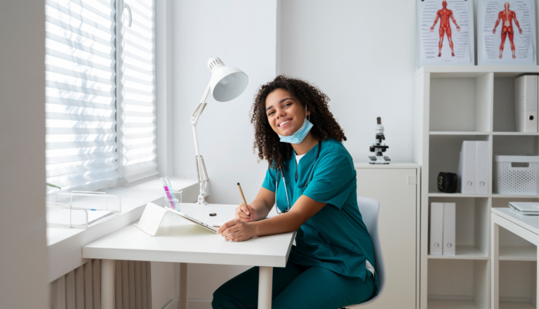 Profissional da área da saúde, vestindo uniforme verde e estetoscópio, sorri enquanto faz anotações em uma mesa dentro de um consultório.