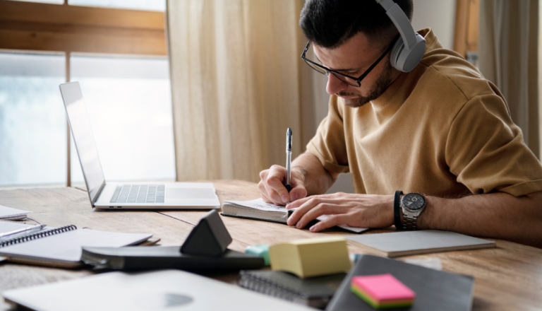 Pessoa usando fones de ouvido e óculos faz anotações em um caderno enquanto utiliza um notebook em uma mesa de trabalho.