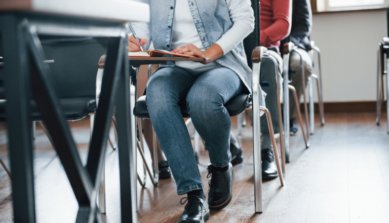 Pessoa sentada em uma sala de aula ou treinamento faz anotações em um caderno, enquanto outras pessoas permanecem sentadas em fileiras ao fundo.