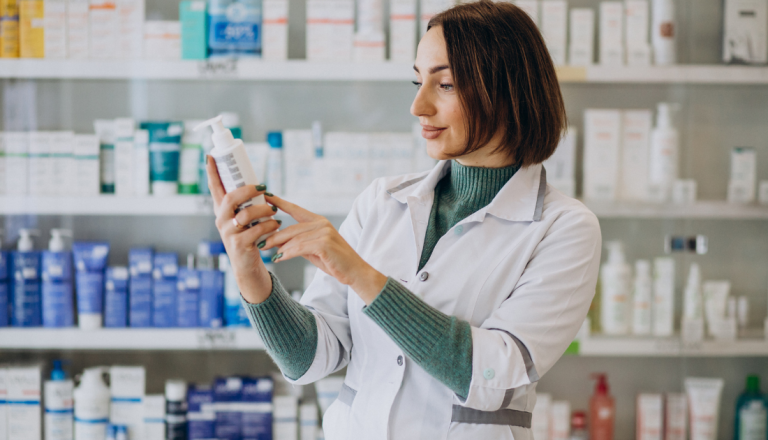 Profissional farmacêutica analisando o rótulo de um produto em uma farmácia, cercada por prateleiras com medicamentos e itens de cuidados pessoais, demonstrando atenção à qualidade e às informações do produto.