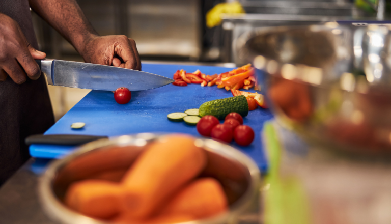 Pessoa corta vegetais sobre uma tábua azul em uma cozinha profissional, com tomates, pepino, pimentão e cenouras dispostos na bancada.