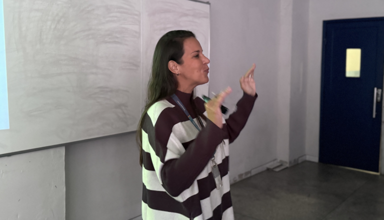 Professora apresenta o projeto CriaEdu3D em uma sala de aula da UNISUAM, gesticulando durante a palestra diante de uma tela de projeção.