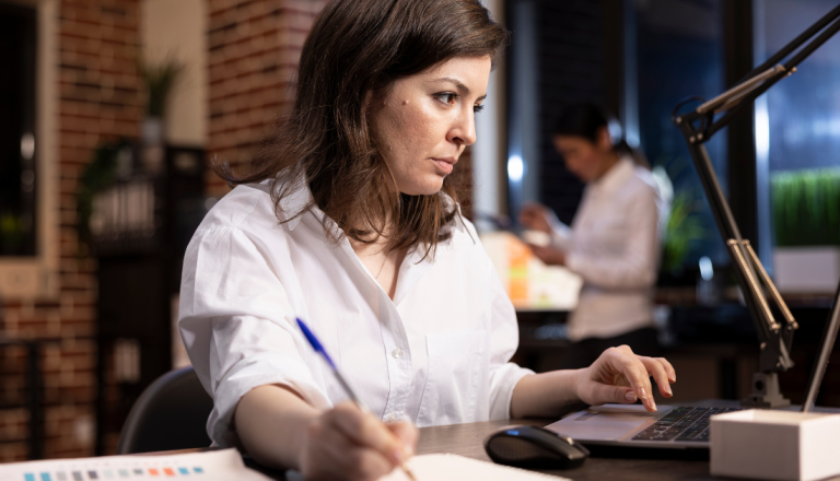 Mulher trabalhando em um escritório, escrevendo em um caderno enquanto utiliza um notebook sobre a mesa. Ao fundo, outra pessoa aparece desfocada no ambiente corporativo.