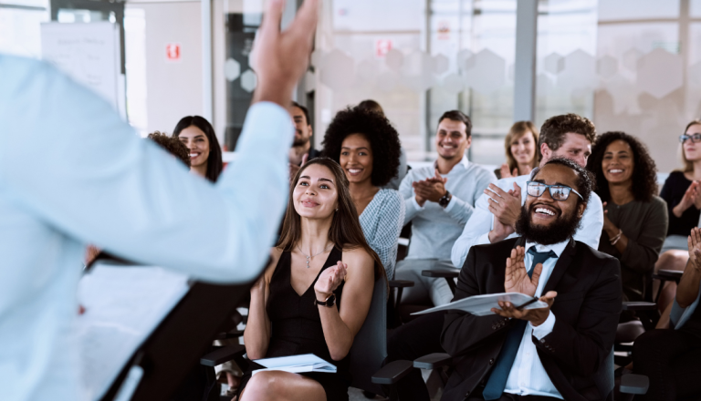 Grupo de pessoas sentadas em uma sala de treinamento ou palestra aplaudindo um palestrante. Os participantes demonstram atenção e entusiasmo durante a apresentação.