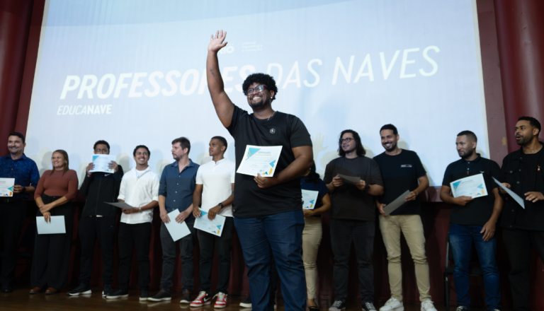 Pessoa segurando um certificado e acenando para o público durante uma cerimônia de certificação em um palco, acompanhada por outras pessoas que também recebem certificados.