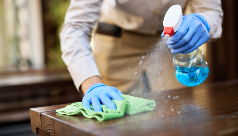 Pessoa utilizando luvas azuis borrifa produto de limpeza sobre uma mesa de madeira enquanto passa um pano de microfibra verde na superfície, realizando a higienização do ambiente.