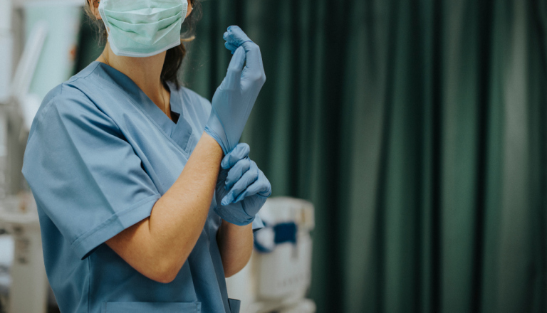 Profissional de saúde usando uniforme cirúrgico azul, máscara facial e luvas descartáveis, ajustando uma das luvas em ambiente hospitalar.