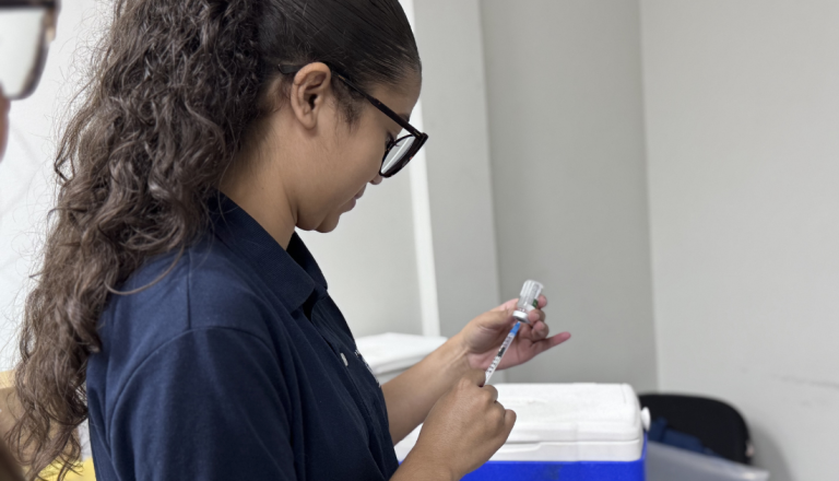 Profissional da área da saúde prepara uma dose de vacina em uma seringa, ao lado de uma caixa térmica utilizada para armazenamento de imunizantes.