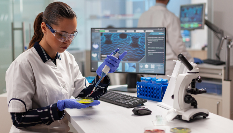 Estudante de Biomedicina realiza análise laboratorial utilizando micropipeta e placa de cultura em laboratório equipado com microscópio e computadores.