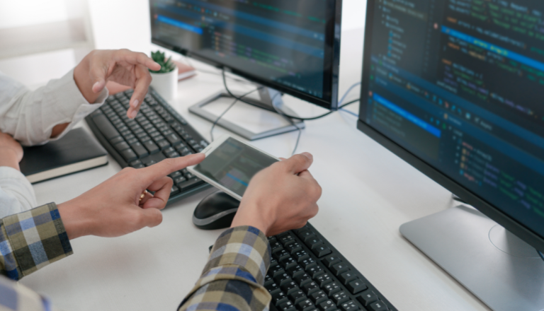 Pessoa utilizando um tablet enquanto analisa códigos de programação exibidos em dois monitores de computador em um ambiente de desenvolvimento de software.