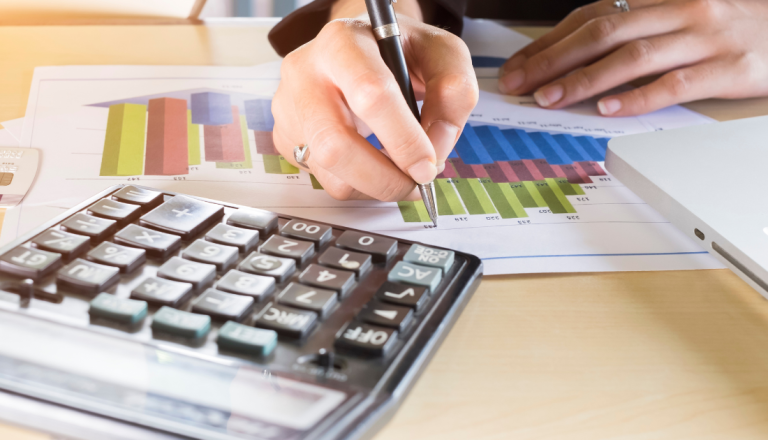 Pessoa analisa gráficos financeiros em uma planilha utilizando calculadora e caneta, com notebook ao lado sobre uma mesa de trabalho.