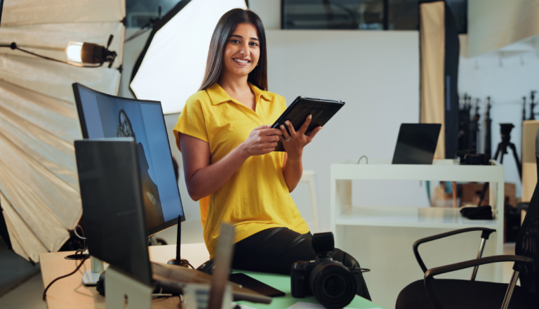 Profissional de publicidade segura um tablet em um estúdio de produção, cercada por equipamentos de fotografia, iluminação e computadores.