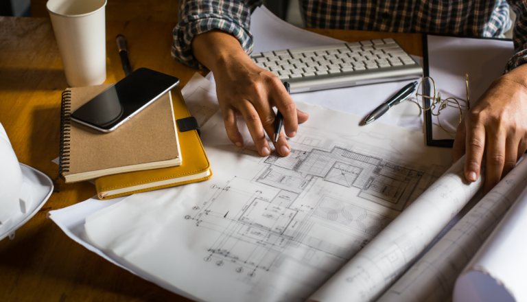 Profissional analisa uma planta arquitetônica sobre uma mesa com computador, caderno, celular e materiais de projeto.