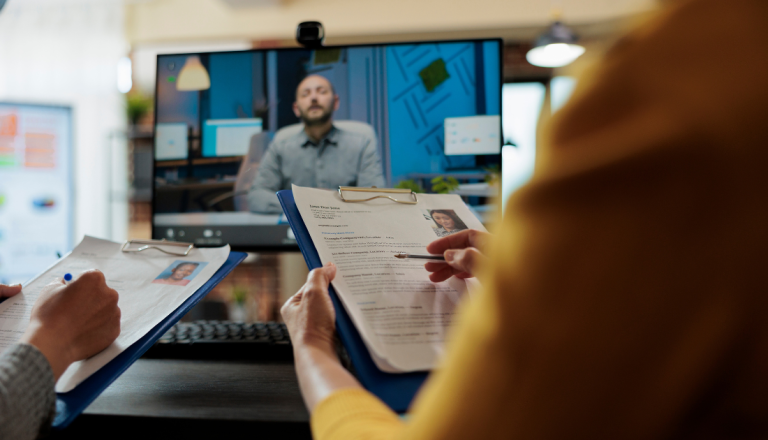 Duas pessoas analisam currículos em pranchetas enquanto participam de uma entrevista de emprego por videoconferência exibida em um monitor.