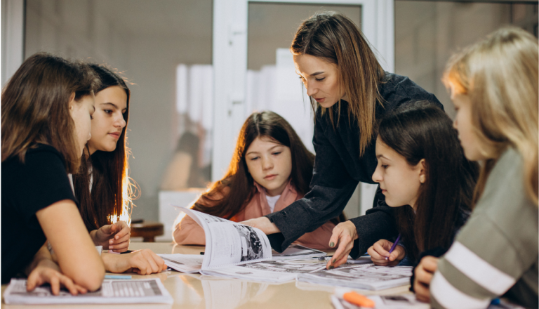 Professora orienta um grupo de estudantes durante uma atividade em sala de aula, apontando o conteúdo de um livro enquanto os alunos acompanham atentamente.
