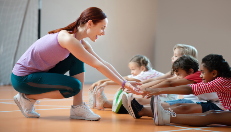 Professora de educação física agachada orienta um grupo de crianças durante um exercício de alongamento em uma quadra esportiva coberta. As crianças estão sentadas no chão, esticando os braços em direção aos pés, enquanto a professora sorri e incentiva a atividade.