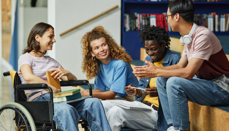 Grupo de quatro estudantes conversa e sorri numa biblioteca. Uma estudante utiliza cadeira de rodas e segura livros no colo, enquanto os restantes colegas estão sentados à sua volta, num ambiente de estudo colaborativo.