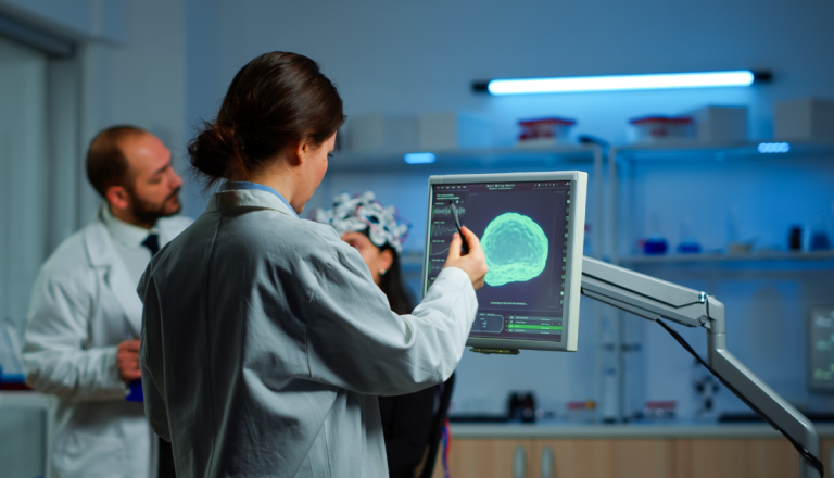 Pesquisadora analisa imagens do cérebro em um monitor durante atividade em laboratório, enquanto outro profissional conversa com uma participante ao fundo.