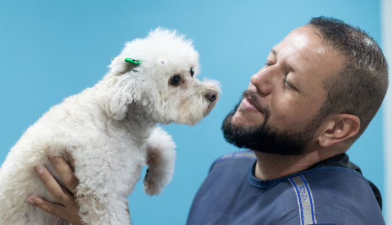 Homem sorri enquanto segura um pequeno cachorro branco nos braços. Os dois se olham de perto, transmitindo uma interação de carinho e confiança. O fundo azul desfocado destaca a conexão entre tutor e pet.