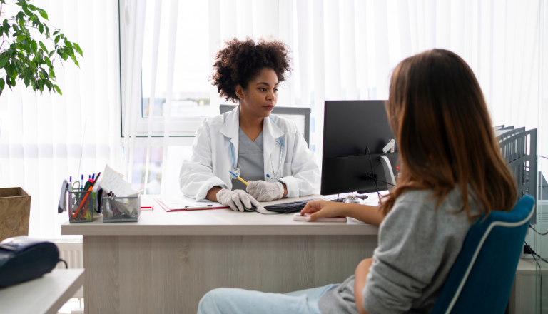 Médica atende uma paciente sentada em consultório durante uma consulta, conversando sobre avaliação e tratamento de saúde.