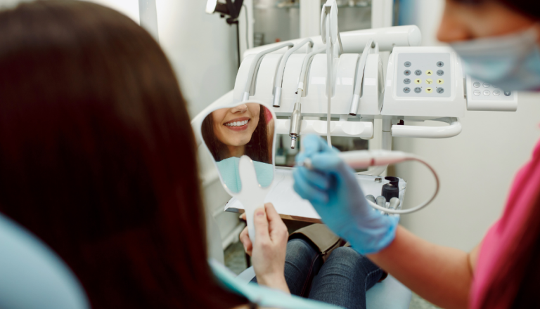 Paciente observa o próprio sorriso em um espelho durante uma consulta odontológica, enquanto uma profissional prepara um equipamento para o atendimento em um consultório moderno.