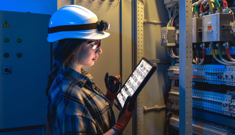 Profissional utilizando capacete, óculos de proteção e luvas inspeciona um painel elétrico com auxílio de um tablet em ambiente industrial.